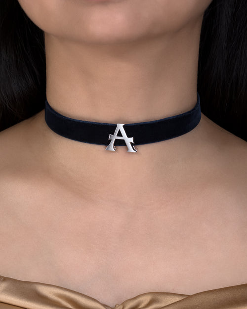Black choker necklace with a silver 'A' pendant on a person wearing a gold garment.